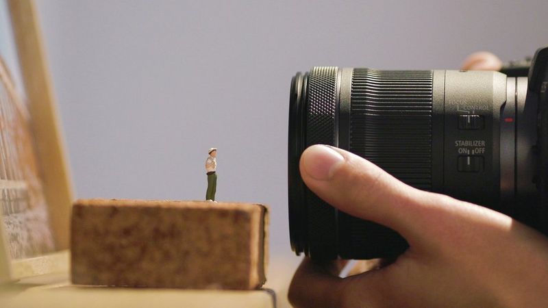 A screenshot from a video explaining centre focus macro. The screenshot shows a user's hand adjusting a Canon lens close to a small model of a human figure.