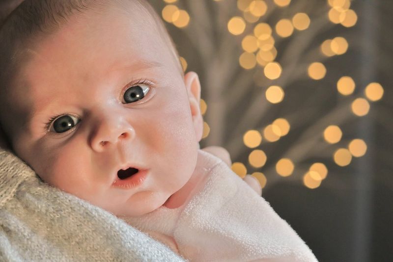 In a photo taken with a Canon EOS R10, a baby looks at the camera over an adult's shoulder, with bokeh lights in the background.