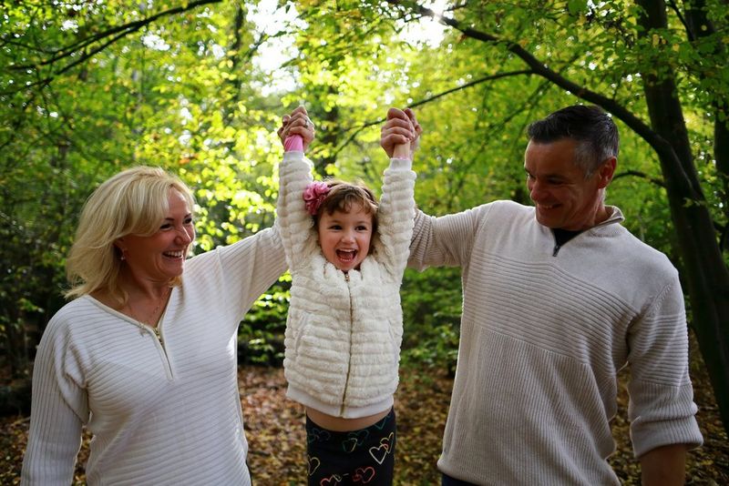 In a photo taken on a Canon EOS RP, a couple raise a smiling child by the arms in a leafy forest. 