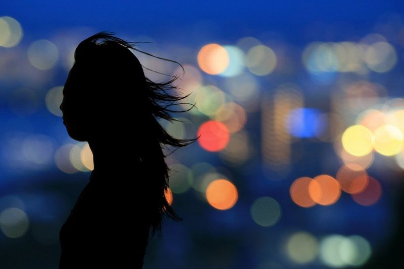 The silhouette of a woman with windblown hair stands against a background of colourful out-of-focus city lights at night.