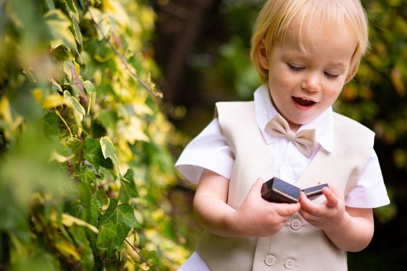 A young boy in a beige-coloured waistcoat, standing next to an ivy covered wall, holds open a ringbox. 