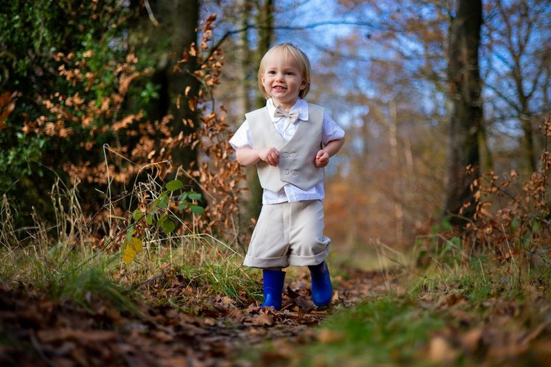 A young boy in a beige waistcoat and bright blue wellies running through a clearing in the woods. 