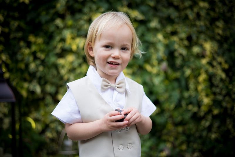 Мальчик в бежевом жилете и галстуке-бабочке стоит перед размытой стеной, увитой плющом, и держит коробочку с кольцом.