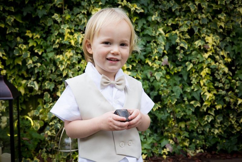 Мальчик в бежевом жилете и галстуке-бабочке стоит перед отчетливо видной стеной, увитой плющом, и держит коробочку с кольцом.