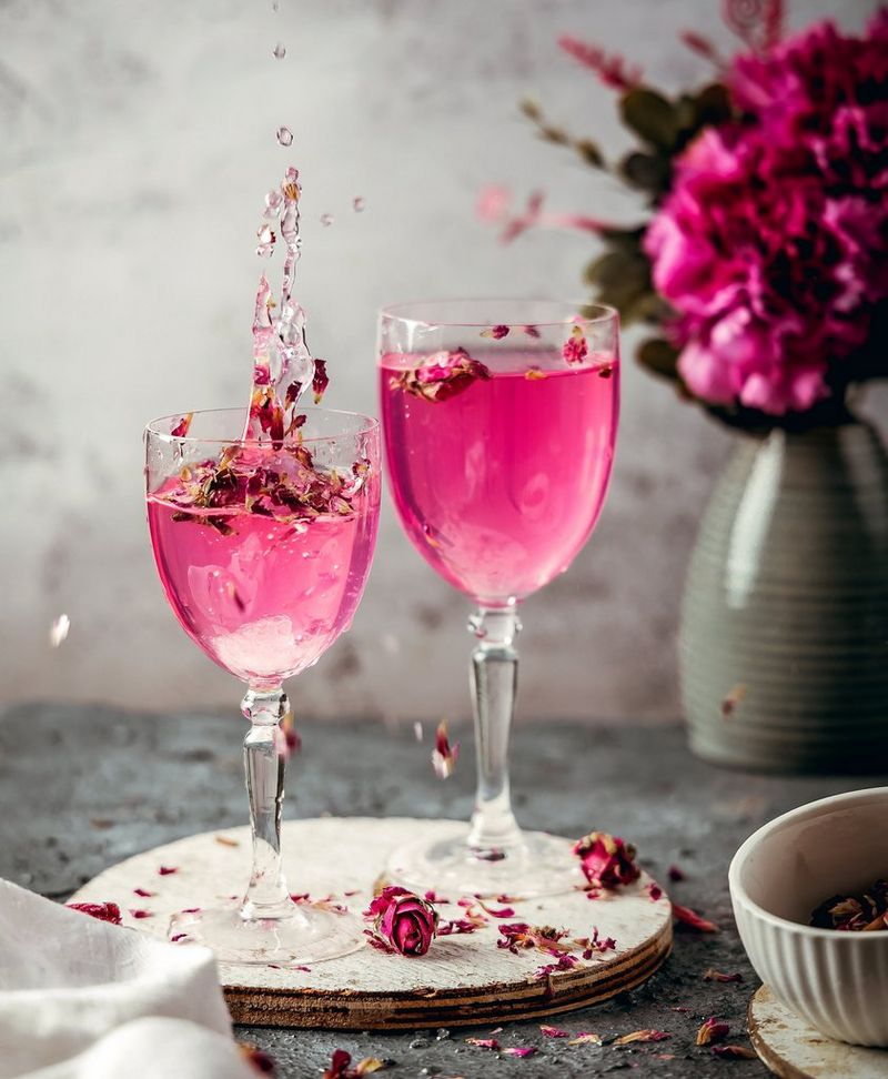Two glasses of bright pink liquid with floating petals in them. The liquid is poured from a height, causing a splash in one glass. 