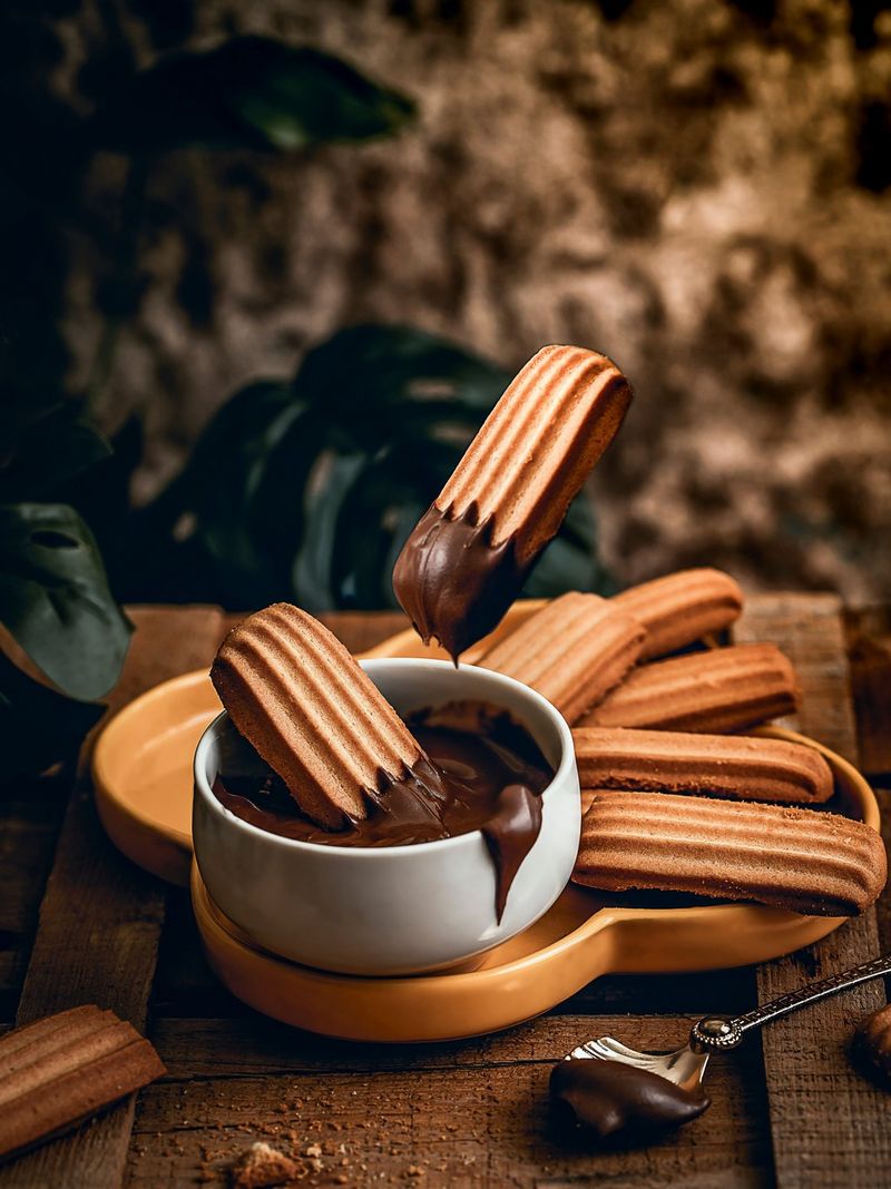 A plate of churros on a table, one dipped in a bowl of chocolate sauce, and one floating above the bowl. 