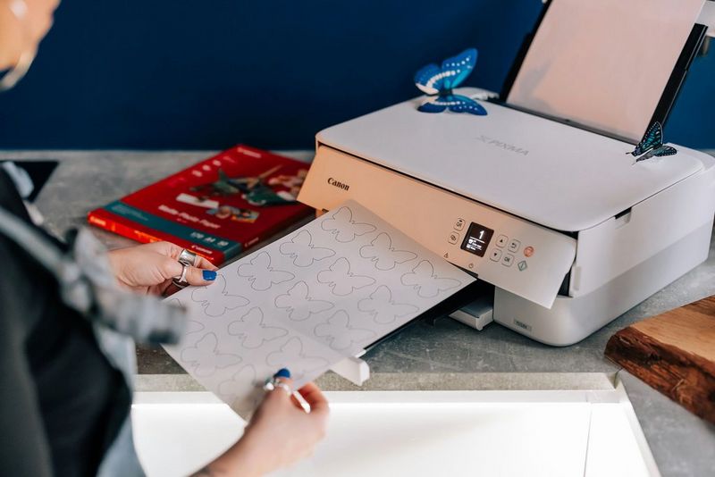 A white Canon printer sits on a desk. In front of it, a woman is holding a printout with a selection of butterfly outlines.