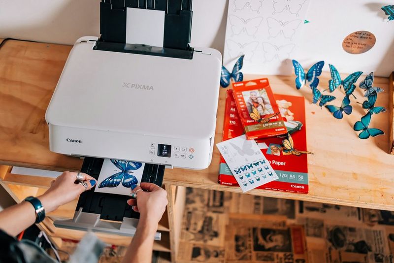 A printer sits on a desk printing a stylised picture of a butterfly, with hands ready to pick it up from the tray. Next to it are several packs of printer paper and colourful butterfly cut-outs. 