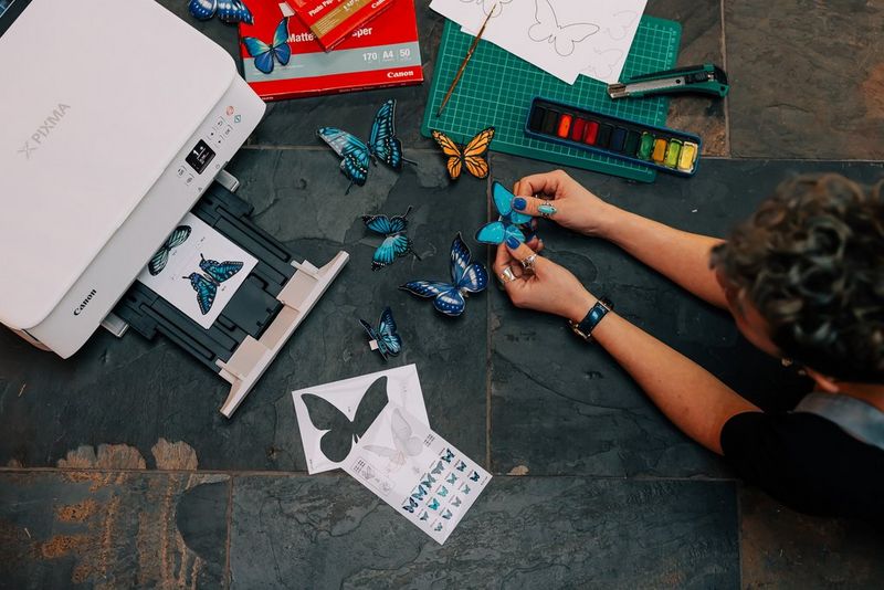Женщина складывает яркие бумажные модели бабочек рядом с принтером серии Canon PIXMA TS5340.