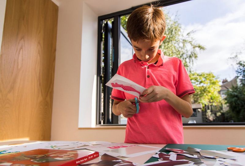 A child stands by a table and uses a pair of scissors to cut a dinosaur papercraft template downloaded from Canon Creative Park.