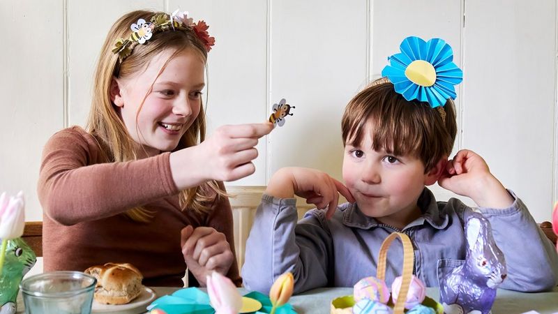 A girl plays with a papercraft bee, while a boy wearing a blue paper flower crown sits next to her. Both crafts were created using a Canon printer and templates from Canon Creative Park. 