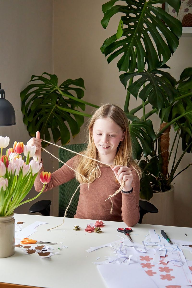 A young girl smiles as she wraps brown string around a metal headband to form the base of a flower crown. 