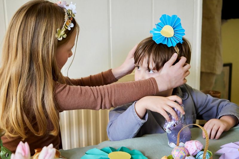 A young girl helps a young boy put a blue paper flower crown on his head