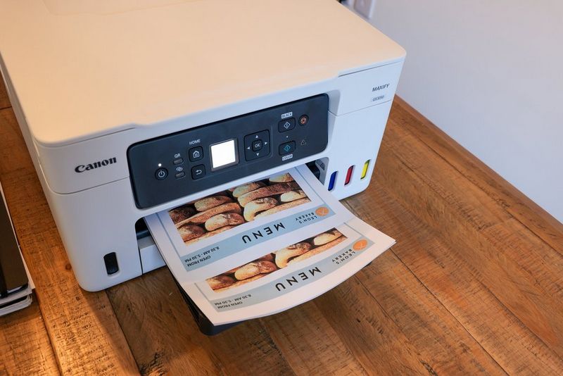 Принтер Canon MAXIFY GX3050 на деревянном столе печатает большое количество меню, созданных в Canon PosterArtist.