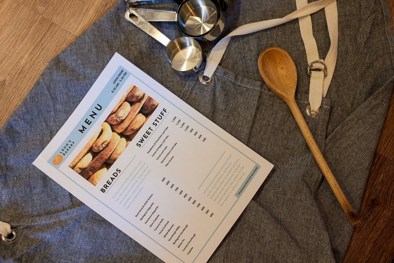 Печатное меню для пекарни, созданное в Canon PosterArtist, лежит на сером фартуке рядом с деревянной ложкой и мерными емкостями для выпечки.