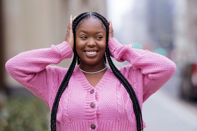 אישה עם צמות ארוכות, לבושה בעליונית ורודה, עומדת בתנוחה כשידיה בשני צדי ראשה.