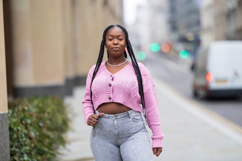 דיוקן של אישה עם צמות ארוכות, לבושה בעליונית ורודה ומכנסי ג'ינס אפורים ברחוב עירוני.