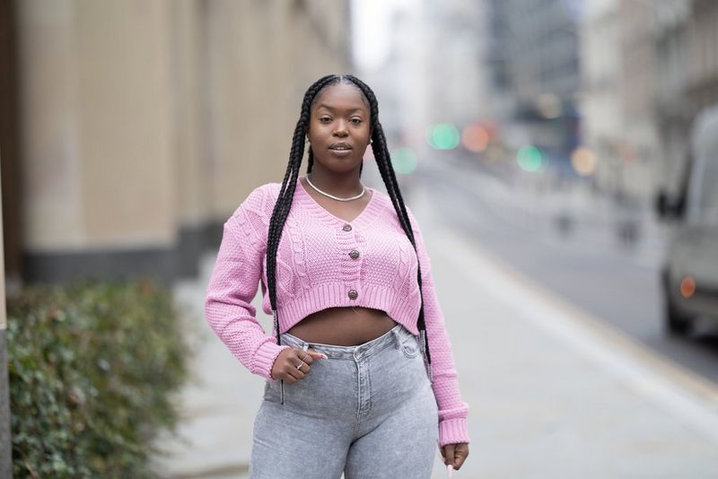 דיוקן במראה קריר יותר של אישה עם צמות ארוכות, לבושה בעליונית ורודה ומכנסי ג'ינס אפורים ברחוב עירוני.