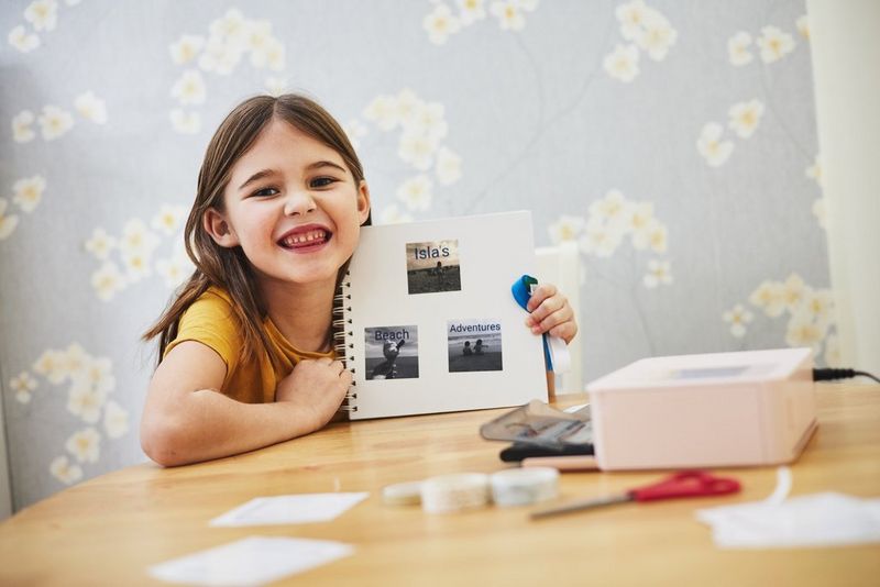 A child smiles at the camera while holding up a handmade storybook, created using a Canon SELPHY CP1500 printer and the Canon SELPHY Photo Layout app.