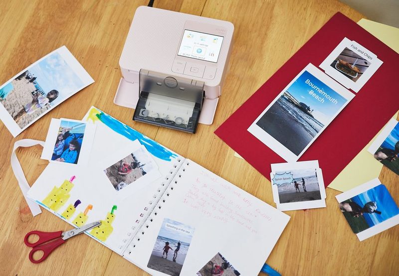 Various prints, a pair of scissors and an open scrapbook next to a pink Canon SELPHY CP1500 lie on a wooden table.