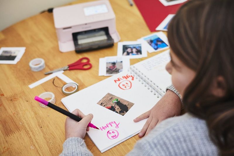 A child uses a coloured pen to write "happy" and draw a smiley face in a scrapbook while various prints, stationery and a Canon SELPHY CP1500 sit on the table.