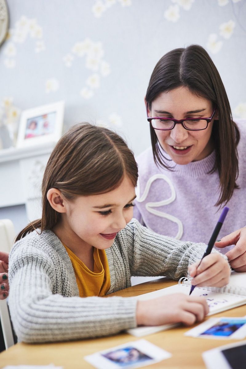 A smiling child uses a pen to write in a scrapbook guided by the adult sat beside her.