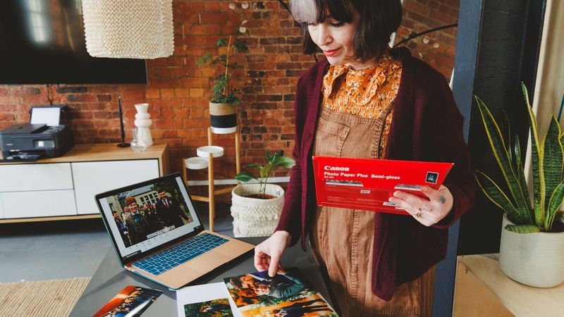 אישה שמחזיקה אריזה של נייר מדפסת של Canon ביחד אחת, ובודקת כמה תדפיסי תמונות שמונחים על השולחן לפניה, כשמחשב נייד מונח לצידם. מדפסות Canon מופיעה על ארונית ברקע.