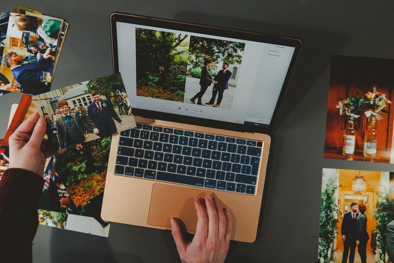 A person looking at wedding images on a laptop, while also holding a photograph from the same wedding in their hand. On the desk around the laptop are several more prints.