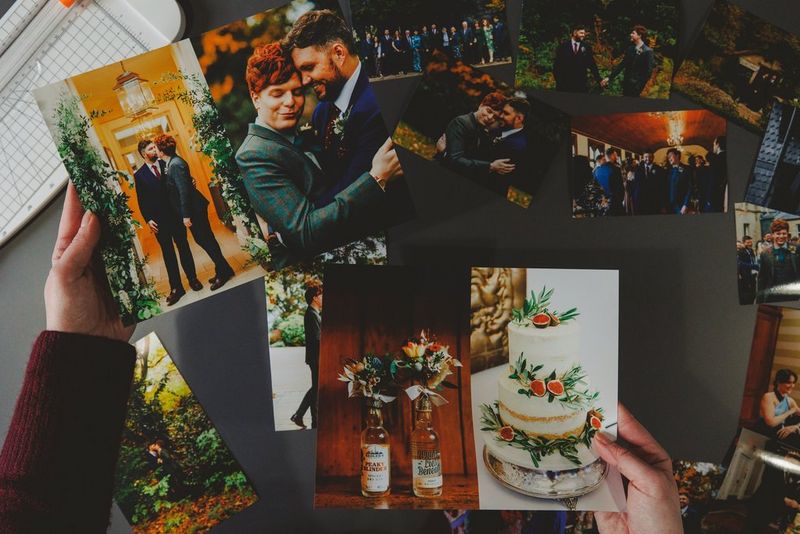 A pair of hands holds up a selection of images from a wedding day including one of the two grooms embracing and a three-tier wedding cake.