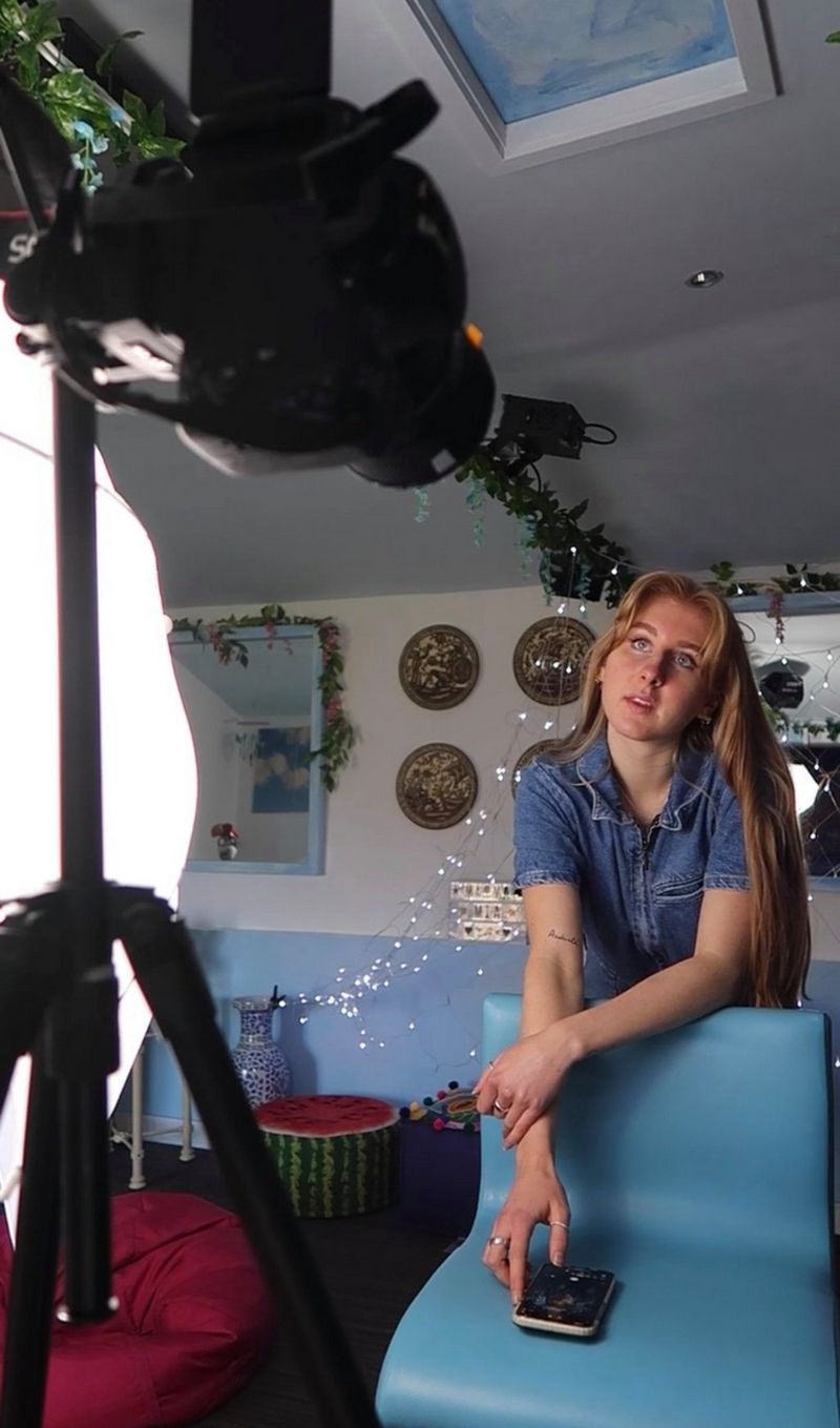 A woman looking at a light in the foreground, leaning over a chair, holding a smartphone.
