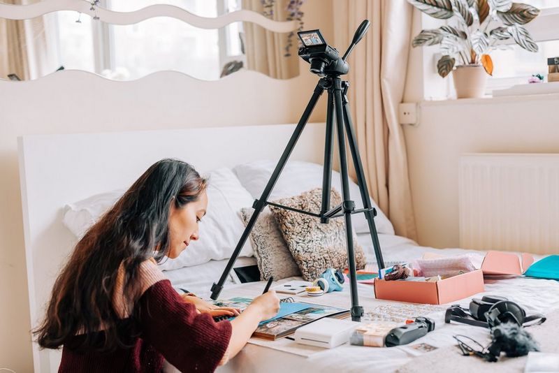 Een naar beneden gerichte Canon PowerShot G7 X Mark III-camera op een statief filmt een vrouw die op een bed een pagina van een scrapbook aan het maken is.