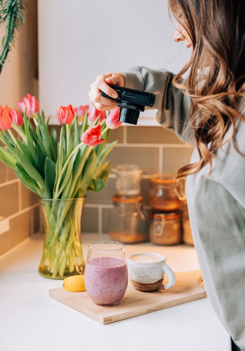 Een jonge vrouw in een grijze trui neemt van bovenaf een foto van een fruitshake en een koffie met schuim.