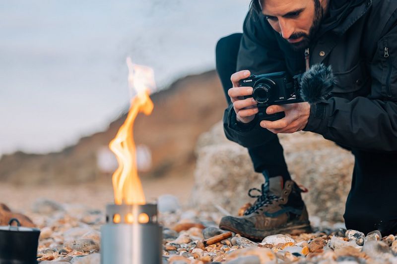 Een man zit op zijn hurken op een strand en fotografeert een kampvuur op een gasbrander met een Canon PowerShot G7 X Mark III.