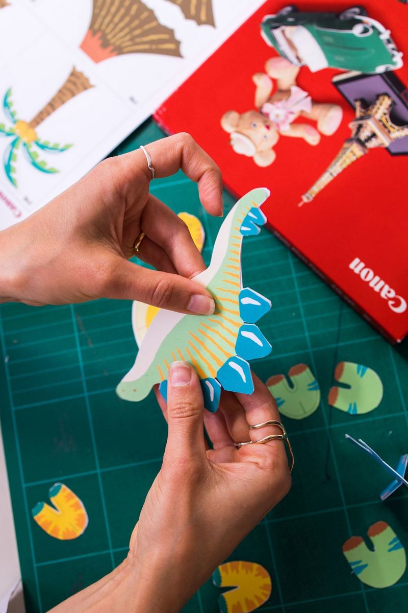 Le mani di un adulto costruiscono un dinosauro cartaceo di Canon Creative Park stampato con una stampante Canon PIXMA, dietro si vede una confezione di carta fotografica Canon.