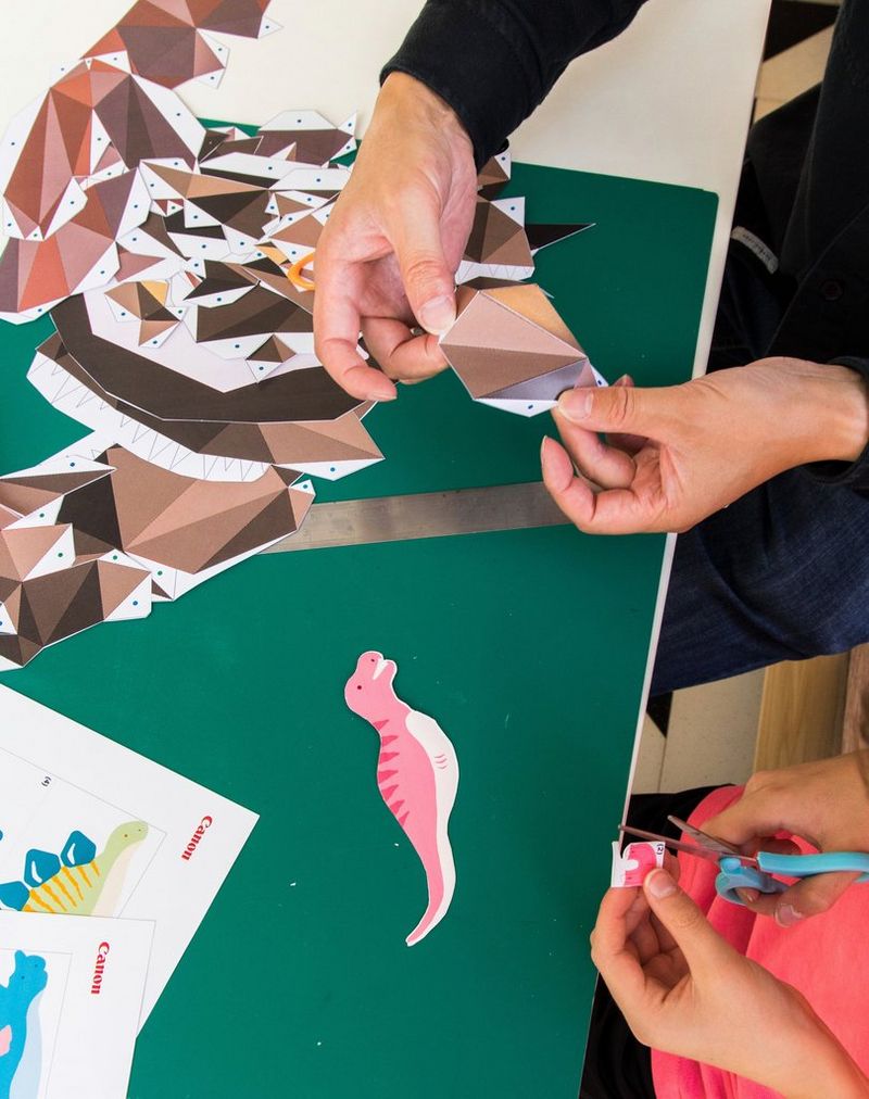 Le mani di un adulto costruiscono la testa di un T-Rex mentre le mani di un bambino ritagliano un semplice dinosauro origami.