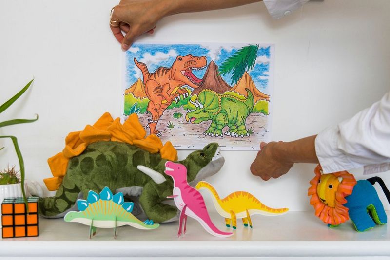 Due mani appendono un disegno colorato di due dinosauri sulla parete dietro a un tavolo con vari modelli di dinosauri di Canon Creative Park.