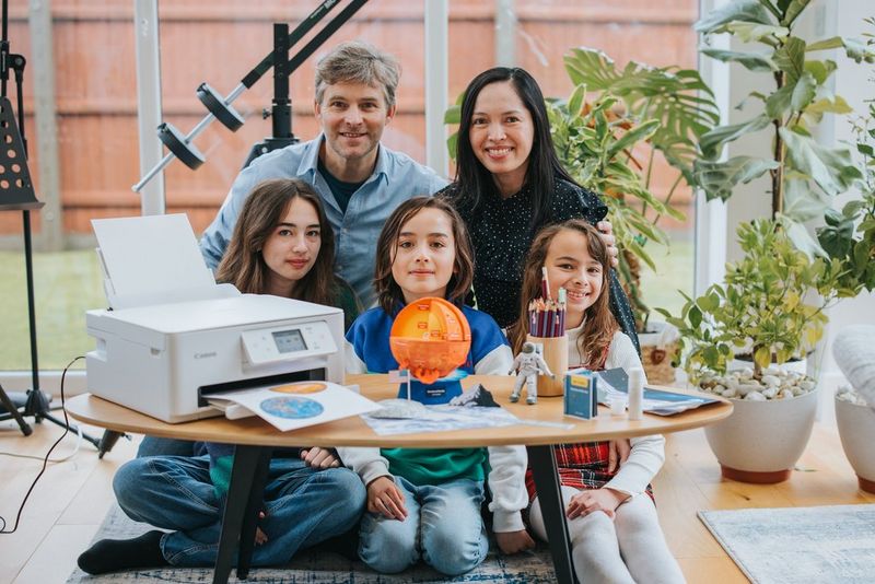 Een gezin dat rond een salontafel zit, met een Canon PIXMA-printer en een selectie papieren modellen met ruimtethema's, gemaakt op basis van Creative Park-sjablonen.