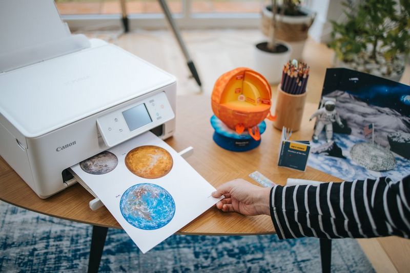 Een hand houdt de rand vast van een Creative Park-sjabloon dat uit een Canon PIXMA-printer komt, die naast de in elkaar gezette papiermodellen met ruimtethema's staat.