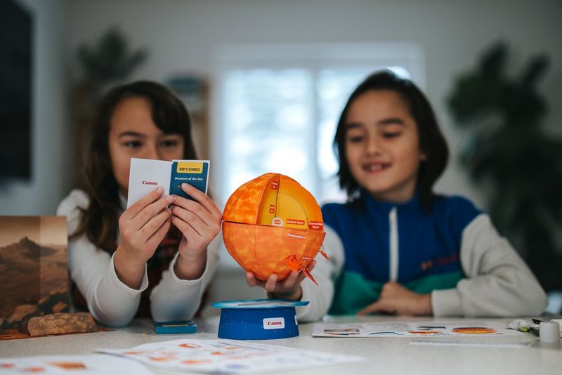 Twee kinderen aan een tafel, één van hen heeft een boek vast met feiten over de zon en het andere kind heeft een in elkaar gezet papieren sjabloon van Creative Park vast.