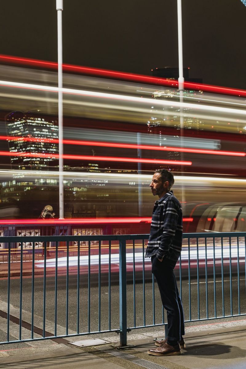 O expunere prelungită a unui bărbat care stă pe trotuar în faţa unor dâre roşii de lumină de la un autobuz care trece.