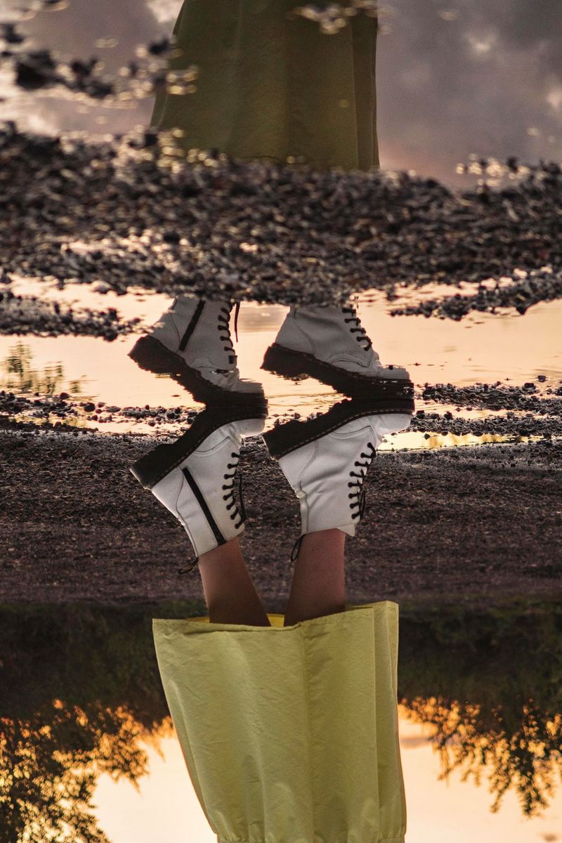 O imagine în oglindă, reflectată într-o baltă, a picioarelor unei femei cu fustă galbenă şi cizme albe, surprinsă de la genunchi în jos.