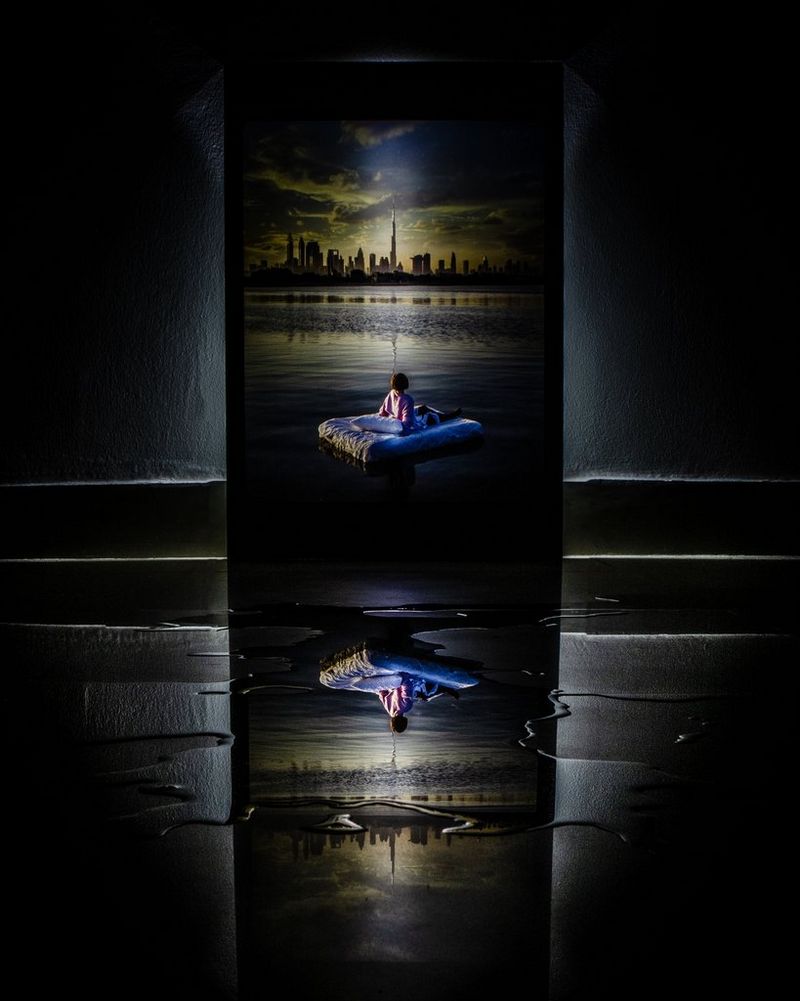 O imagine în oglindă, reflectată într-o baltă de apă, a unui copil care stă pe o saltea plutitoare, cu linia orizontului oraşului în spate.
