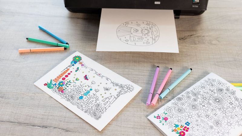 Black Canon PIXMA printer with a colouring printout and two colouring pages and markers on a desk.