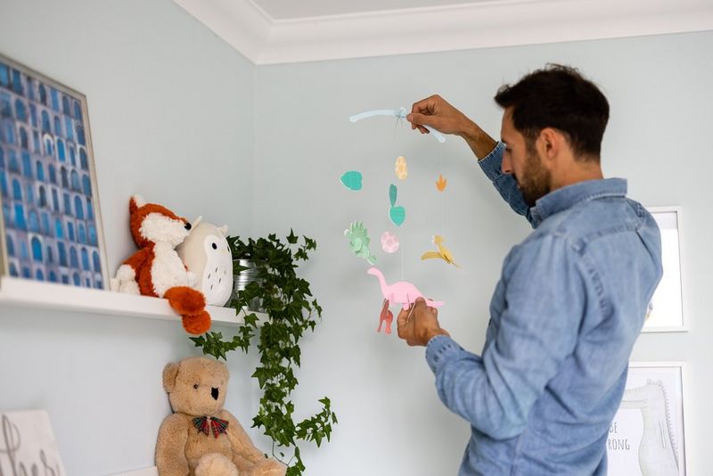 Мужчина вешает бумажный мобиль с динозаврами в детской комнате.