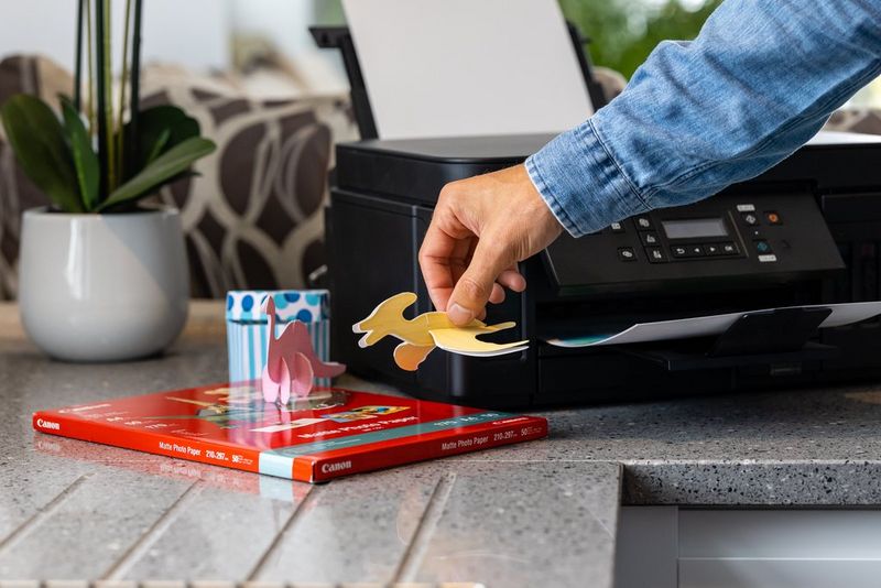 Iemand houdt een papieren dinosaurus vast naast een Canon PIXMA G6050-printer.