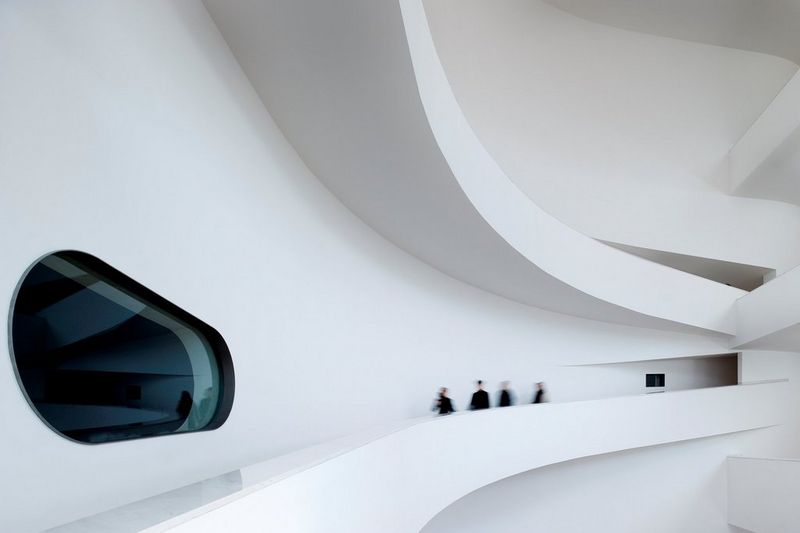 Četiri osobe u zamućenom pokretu dok hodaju velikim prolazom unutar ogromne bijele zgrade s futurističkim prozorima i zidovima.