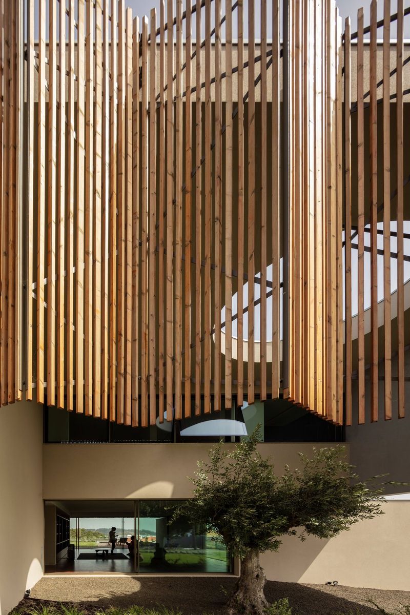 Ogromne drvene letvice tamno krem boje vise okomito s velike zgrade. U prostoriji u prizemlju zgrade naziru se obrisi dvoje ljudi.