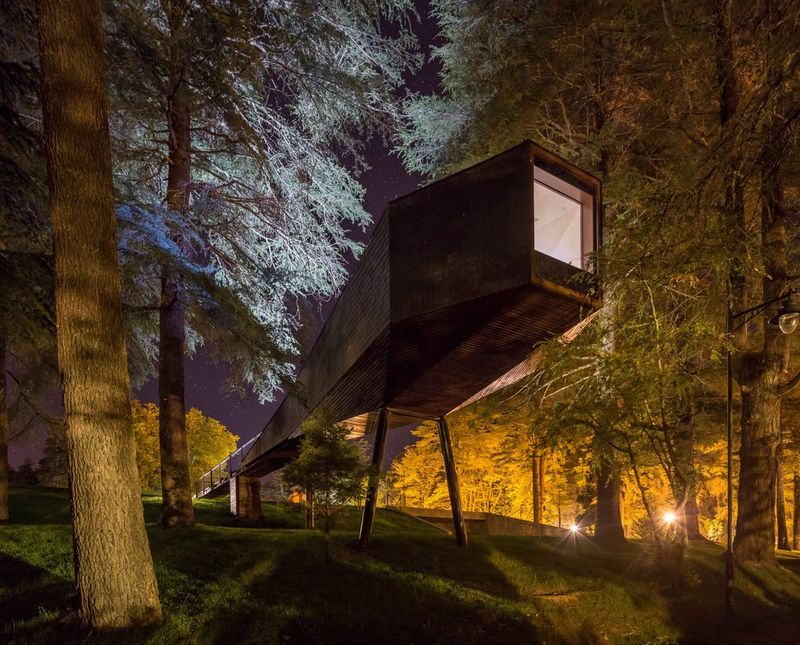 Duguljasta, podignuta drvena zgrada noću. Stabla u okruženju osvijetljena su jarkim svjetlima.