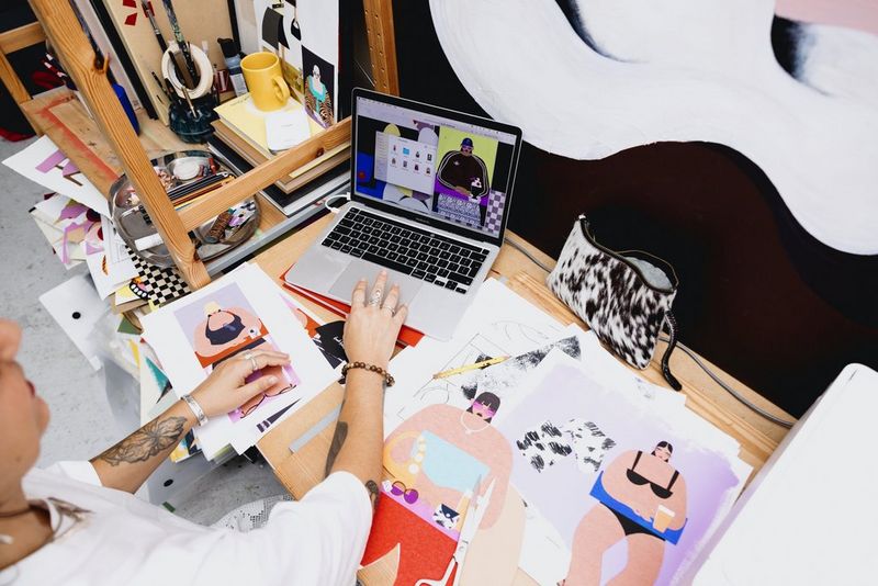 Collagekunstenaar Beth Fraser bekijkt een scan van een van haar vorige werken op haar laptop en verschillende lopende projecten liggen op de tafel voor haar.