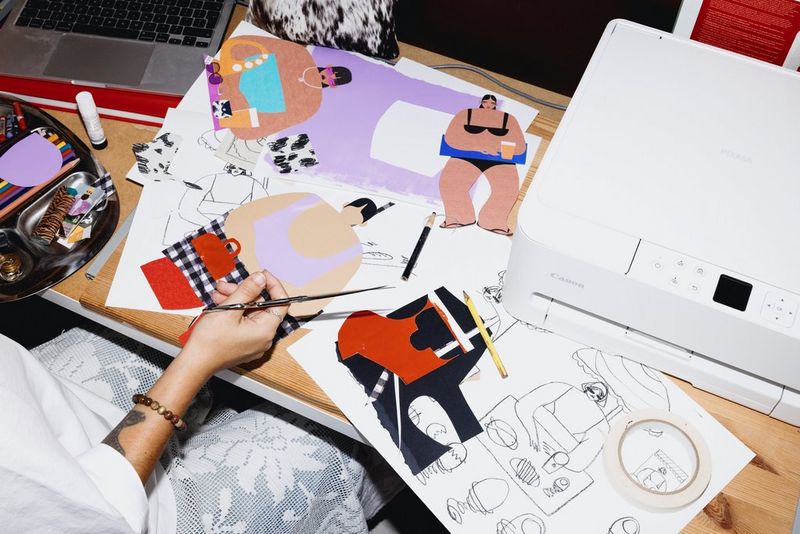 Een bovenaanzicht van de werkbank van collagekunstenaar Beth Fraser, waarop verschillende lopende projecten, een vel met schetsen en een multifunctionele Canon PIXMA-printer liggen.